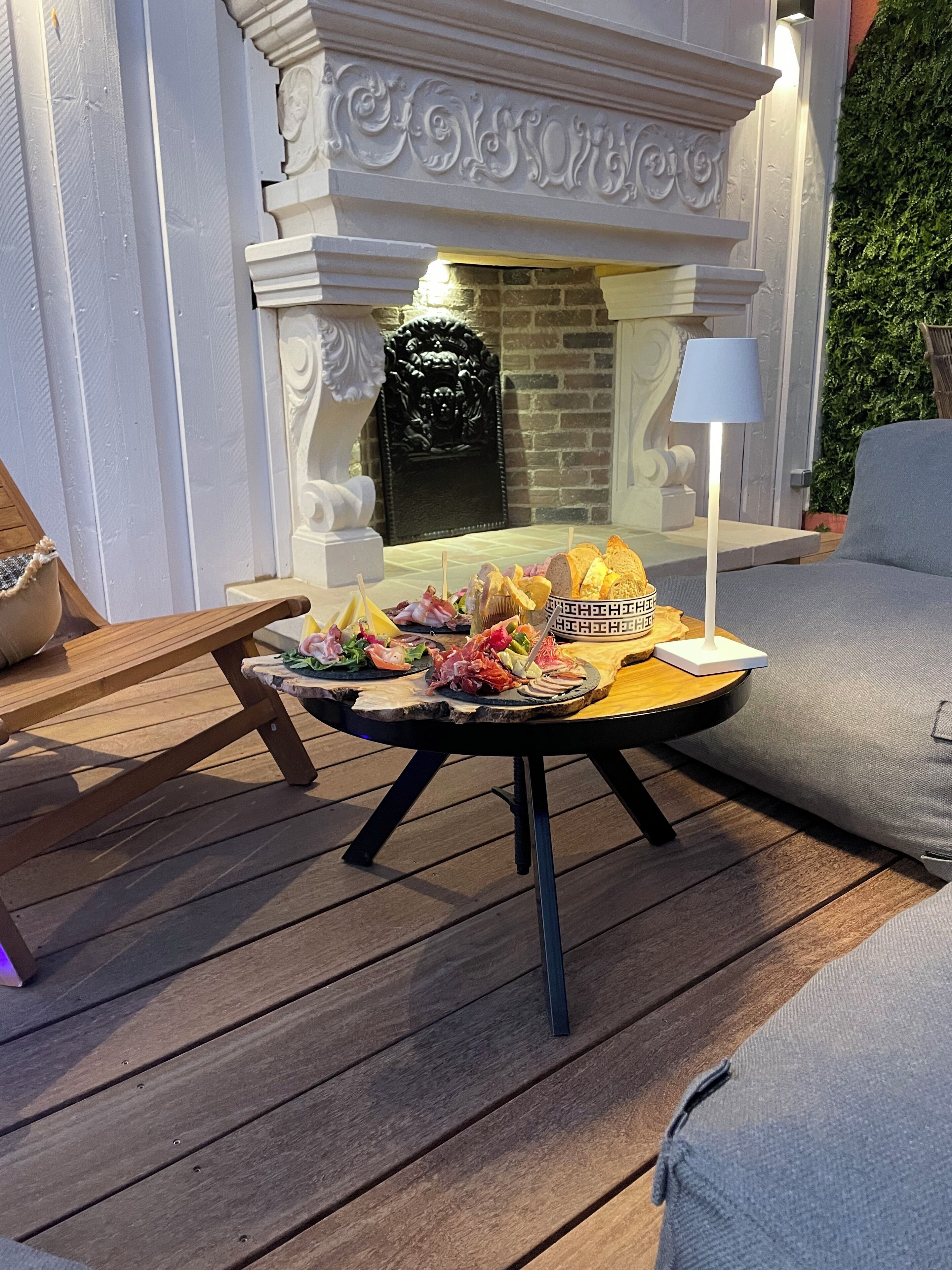 Una terraza de madera con una mesa redonda dispuesta para una comida, una chimenea blanca y un sofá gris.