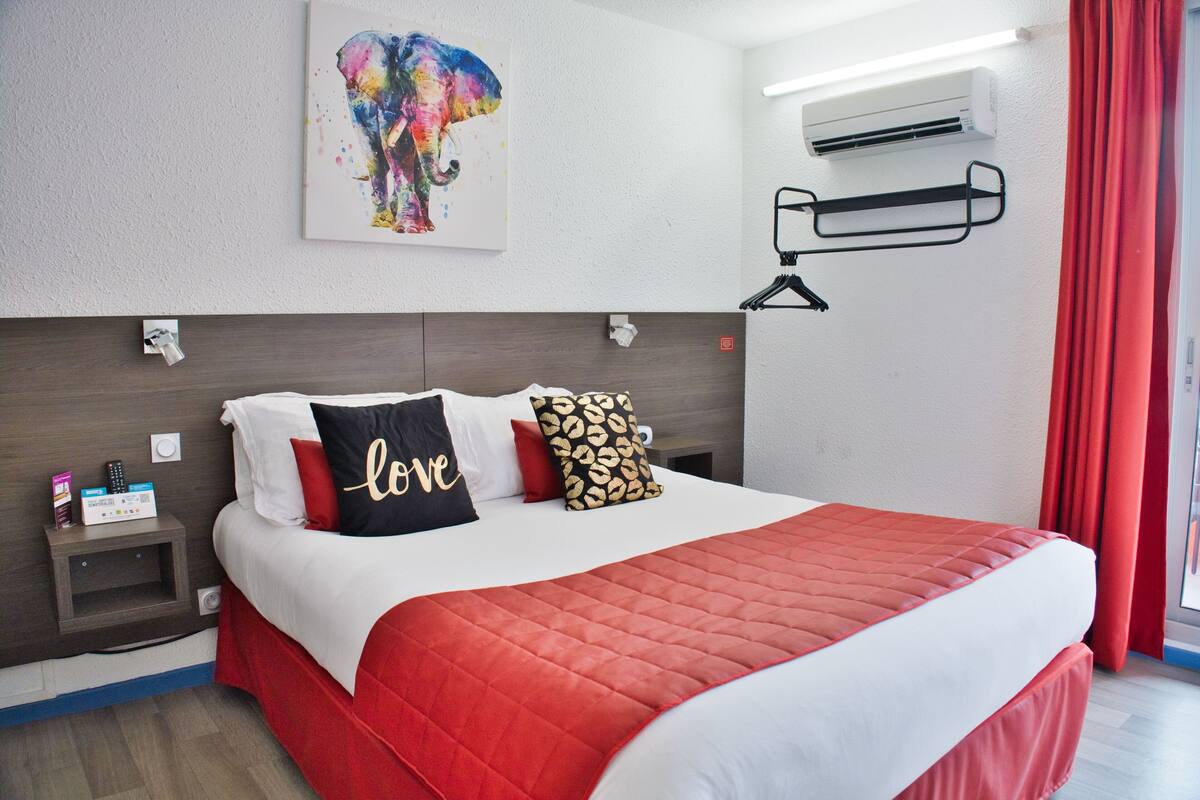 A hotel room with a bed, a nightstand, a wall-mounted air conditioner, and a red curtain.