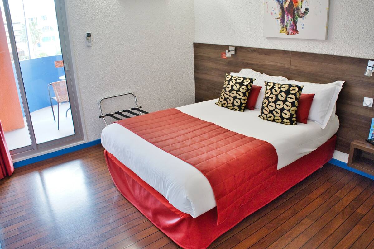 A hotel room with a large bed, a wooden floor, a view of the outside, and a painting on the wall.