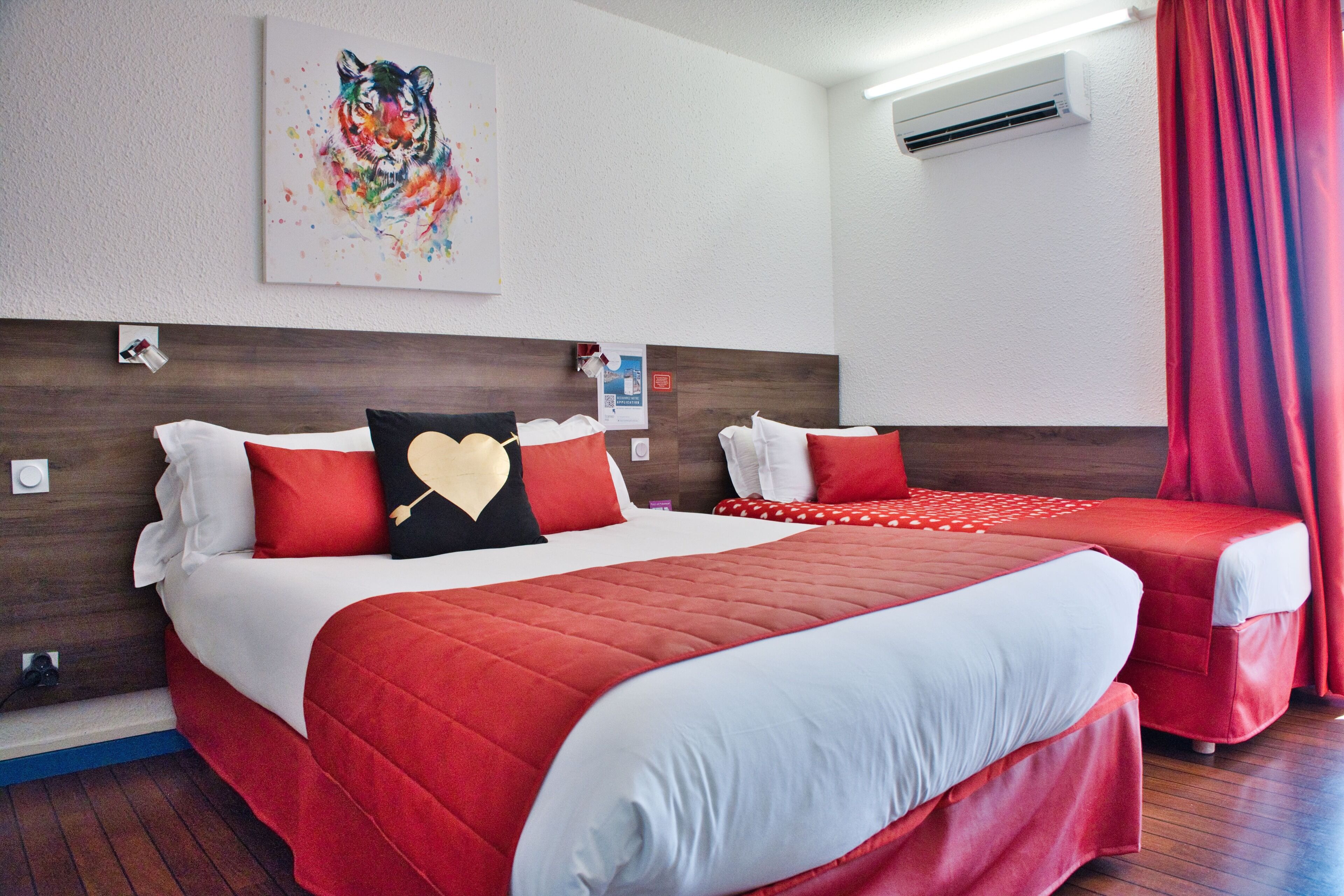 Een hotelkamer met twee bedden, rode beddengoed, een kleurrijke tijgerprint aan de muur en een airconditioner.