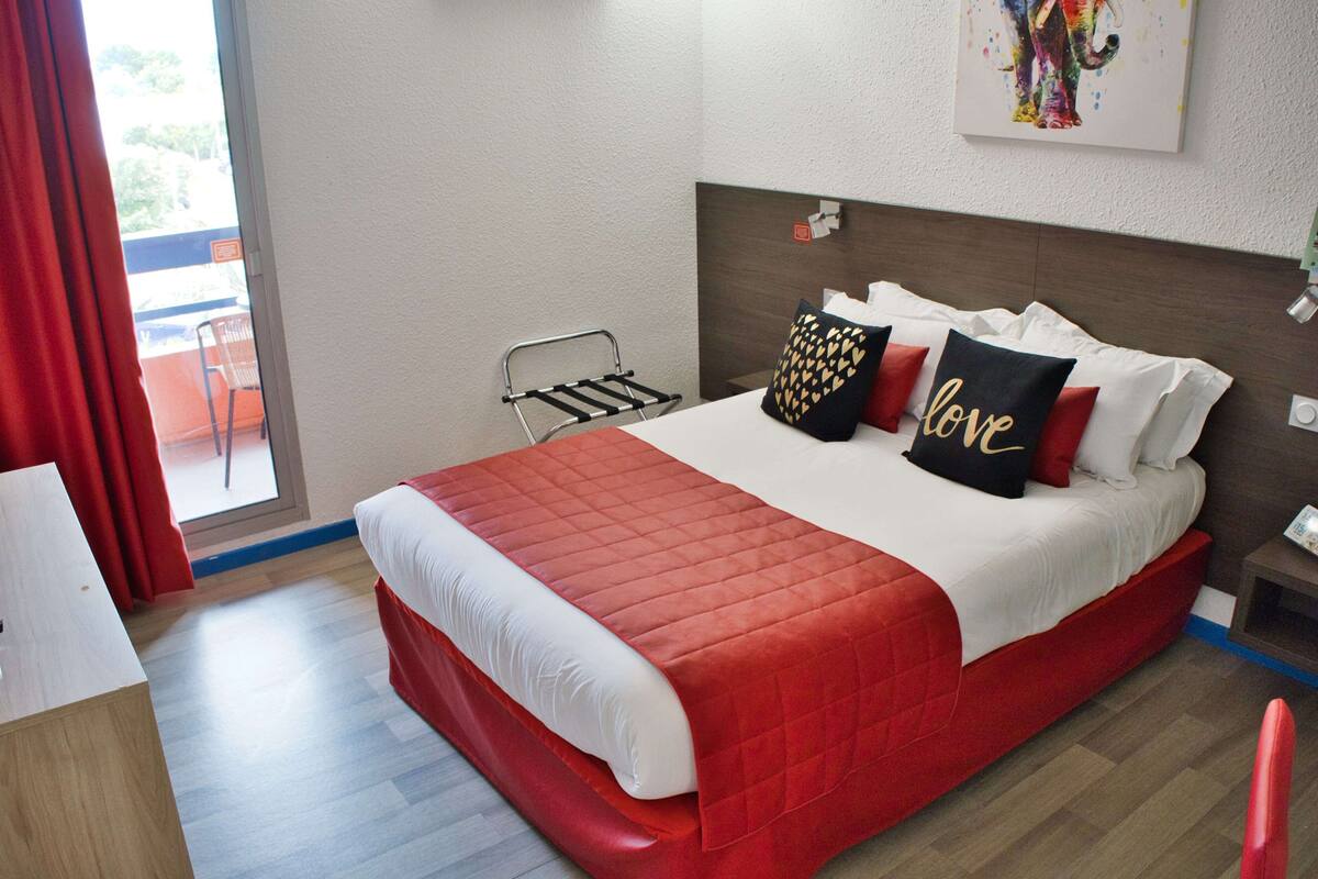 A hotel room with a large bed, a small table, a chair, a window with curtains, and a painting on the wall.