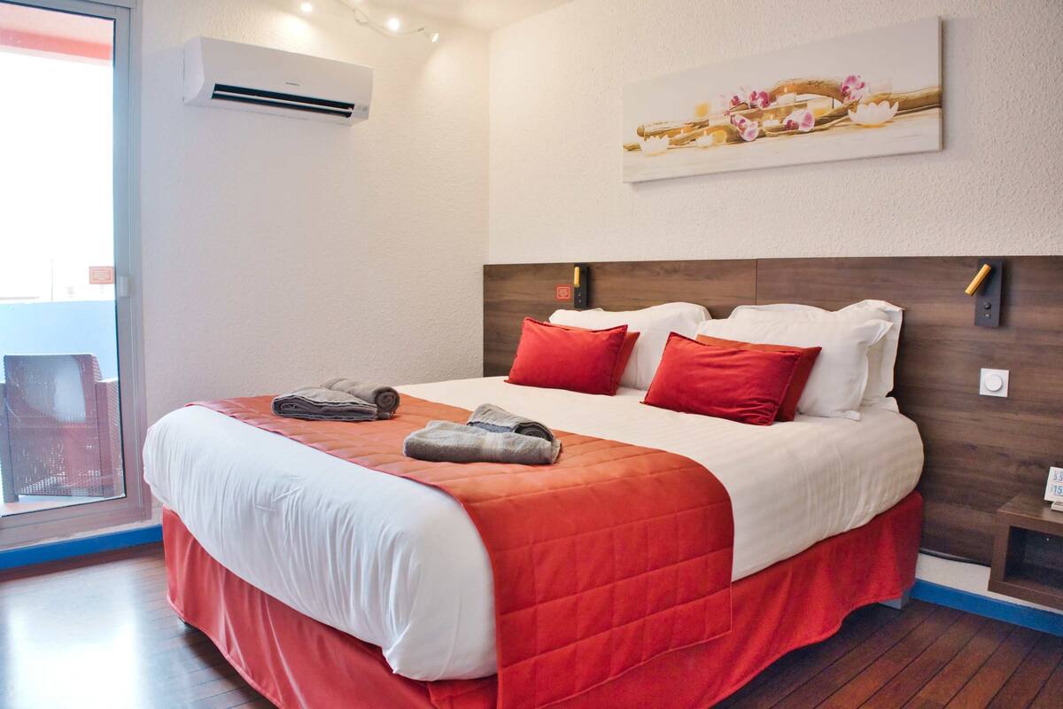 A hotel room with a large bed, red and white bedding, an air conditioning unit, a wooden headboard, a painting on the wall, and a window with a view.