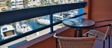 A balcony with wicker chairs and a table, overlooking a marina with boats and buildings.