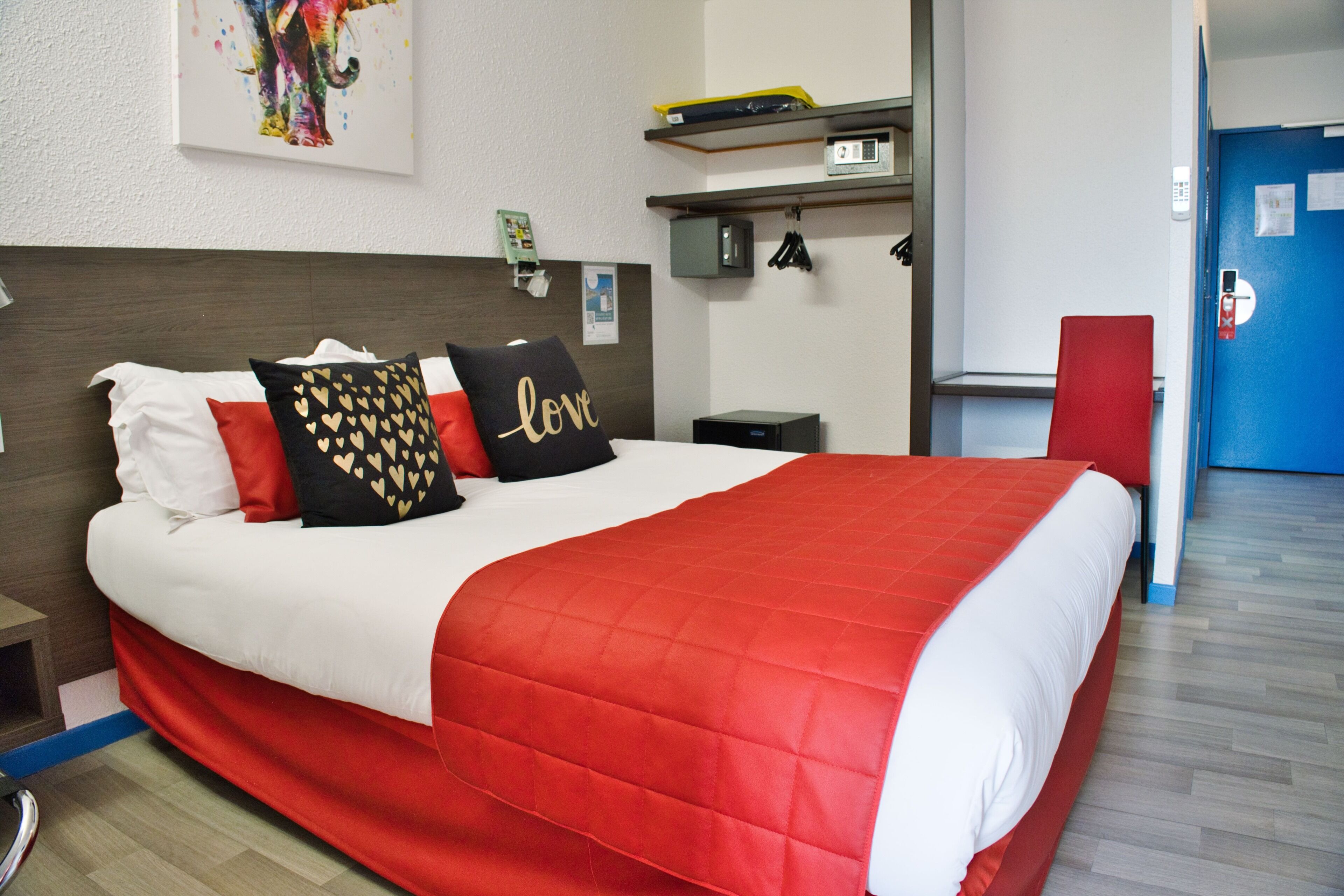 Habitación de hotel con una cama grande, cabecera, mesita de noche, escritorio, silla, estante con objetos, puerta y un cuadro en la pared.