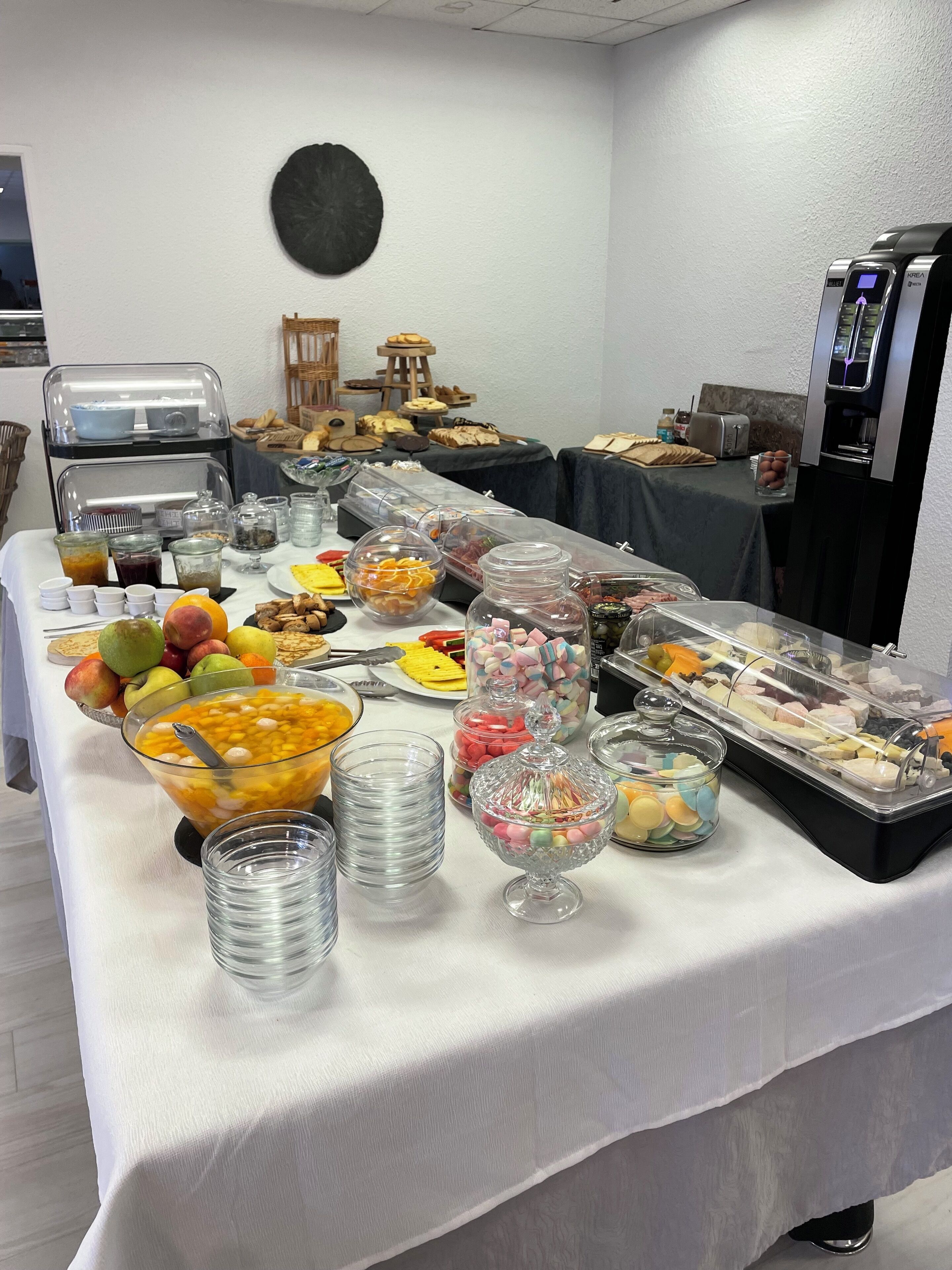 Una mesa de buffet con diversos alimentos y bebidas, incluyendo frutas, pastelería y una máquina de café.