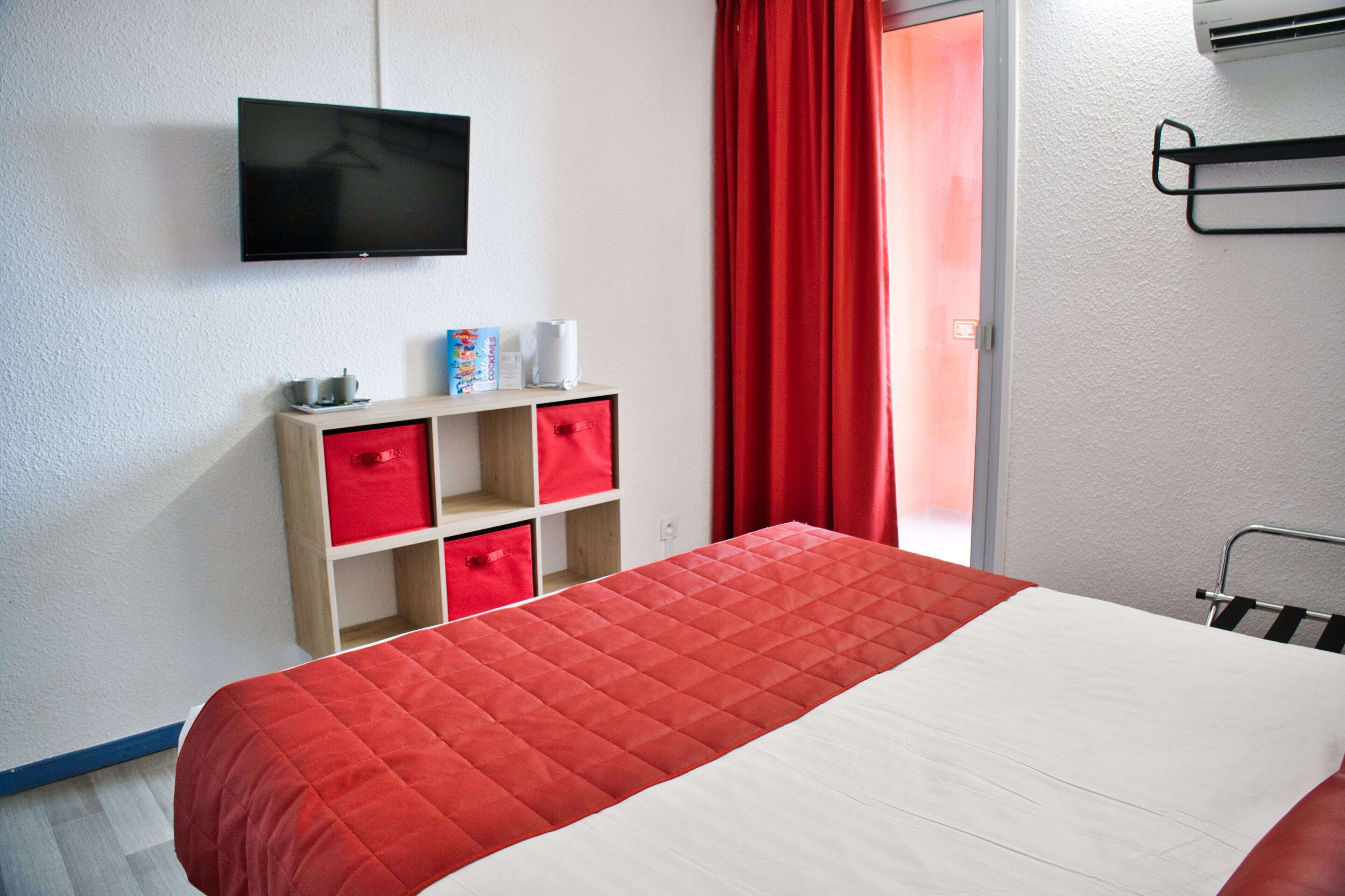 Una habitación de hotel con una cama, un televisor montado en la pared, una estantería con cajas rojas y una ventana con cortinas rojas.
