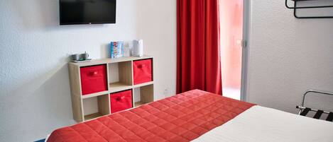 Una habitación de hotel con una cama, un televisor montado en la pared, una estantería con cajas rojas y una ventana con cortinas rojas.
