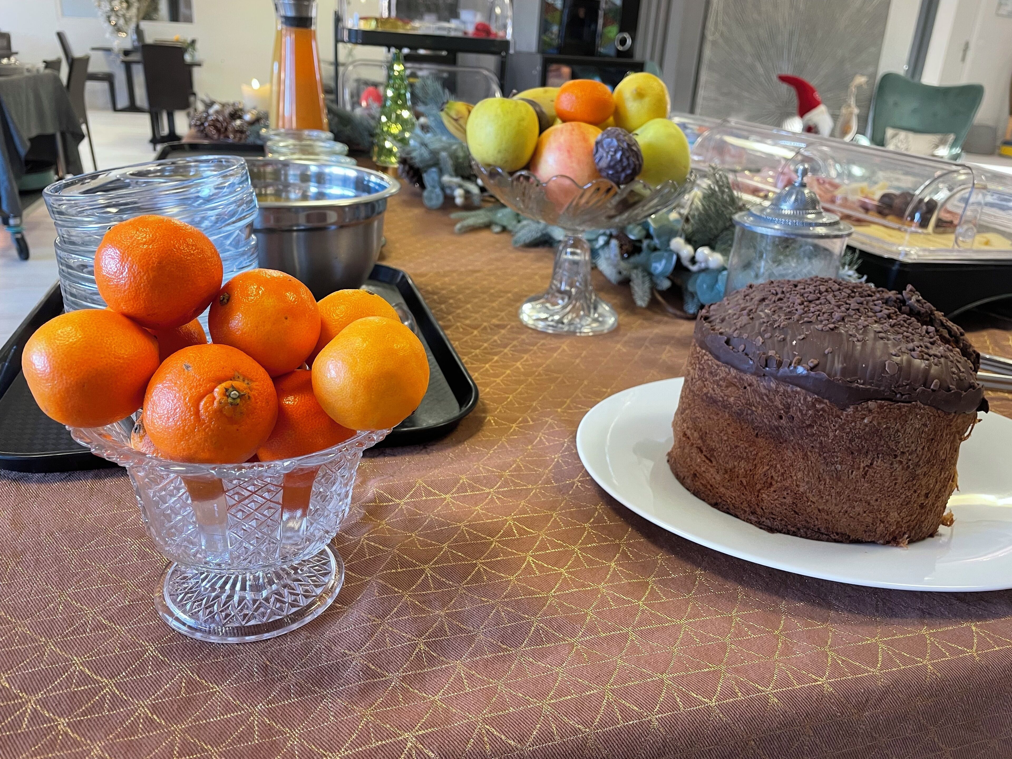 A table with a glass bowl of oranges, a chocolate cake on a plate, and a fruit display with various fruits.