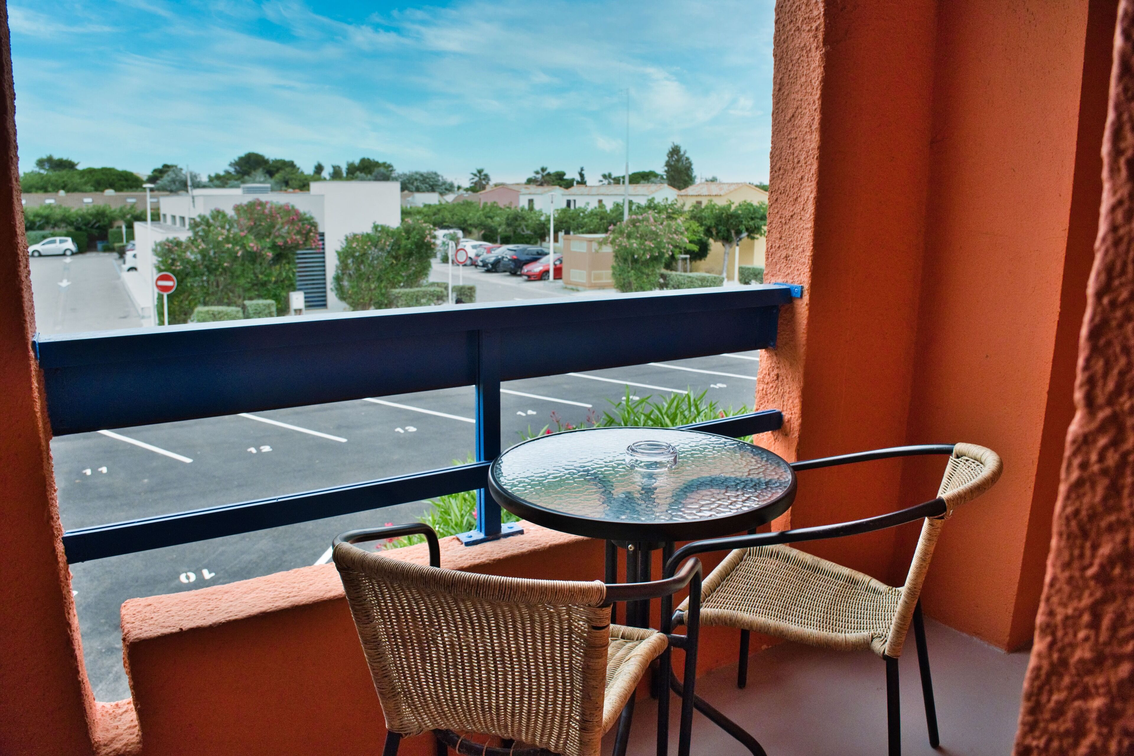 Un balcon avec une table en verre et des chaises en osier, donnant sur un parking et des immeubles.