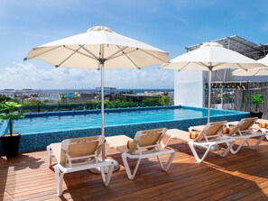 Un espace piscine sur le toit, avec des chaises longues et des parasols, une vue sur l’océan et une piscine carrelée.