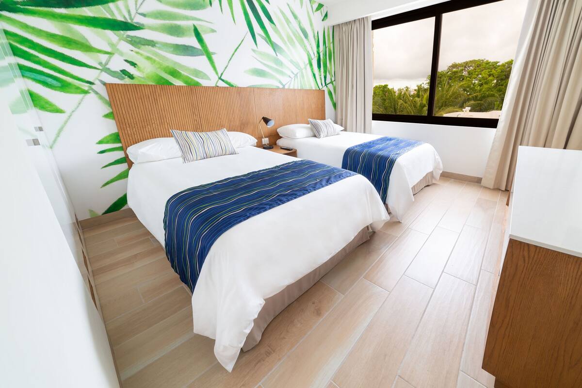 Habitación de hotel con dos camas, un cabecero de madera, un ventanal con vistas y una pared con un estampado de hojas verdes.