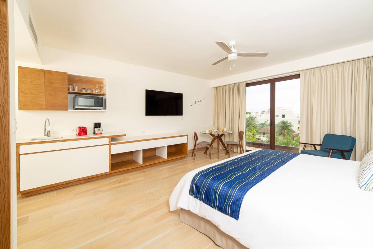 Habitación de hotel con cama, escritorio, televisor, ventana con cortinas y vistas a edificios.