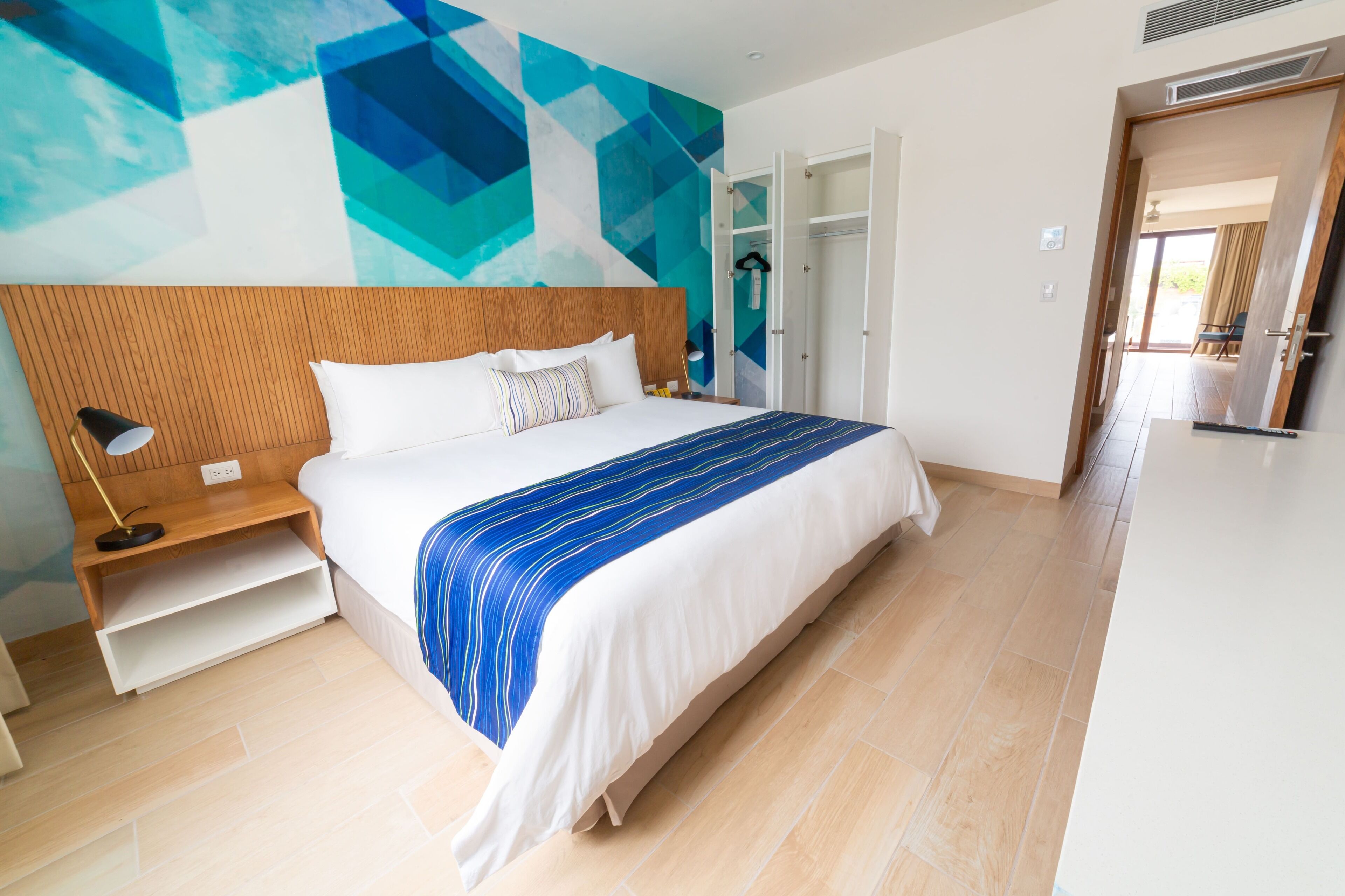 A modern hotel room with a large bed, a wooden headboard, and a geometric wall mural.