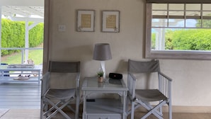Ein Zimmer mit weißer Decke, einem Fenster mit beiger Jalousie, zwei gerahmten Bildern an der Wand, einem kleinen Tisch mit Lampe und einem Stuhl.