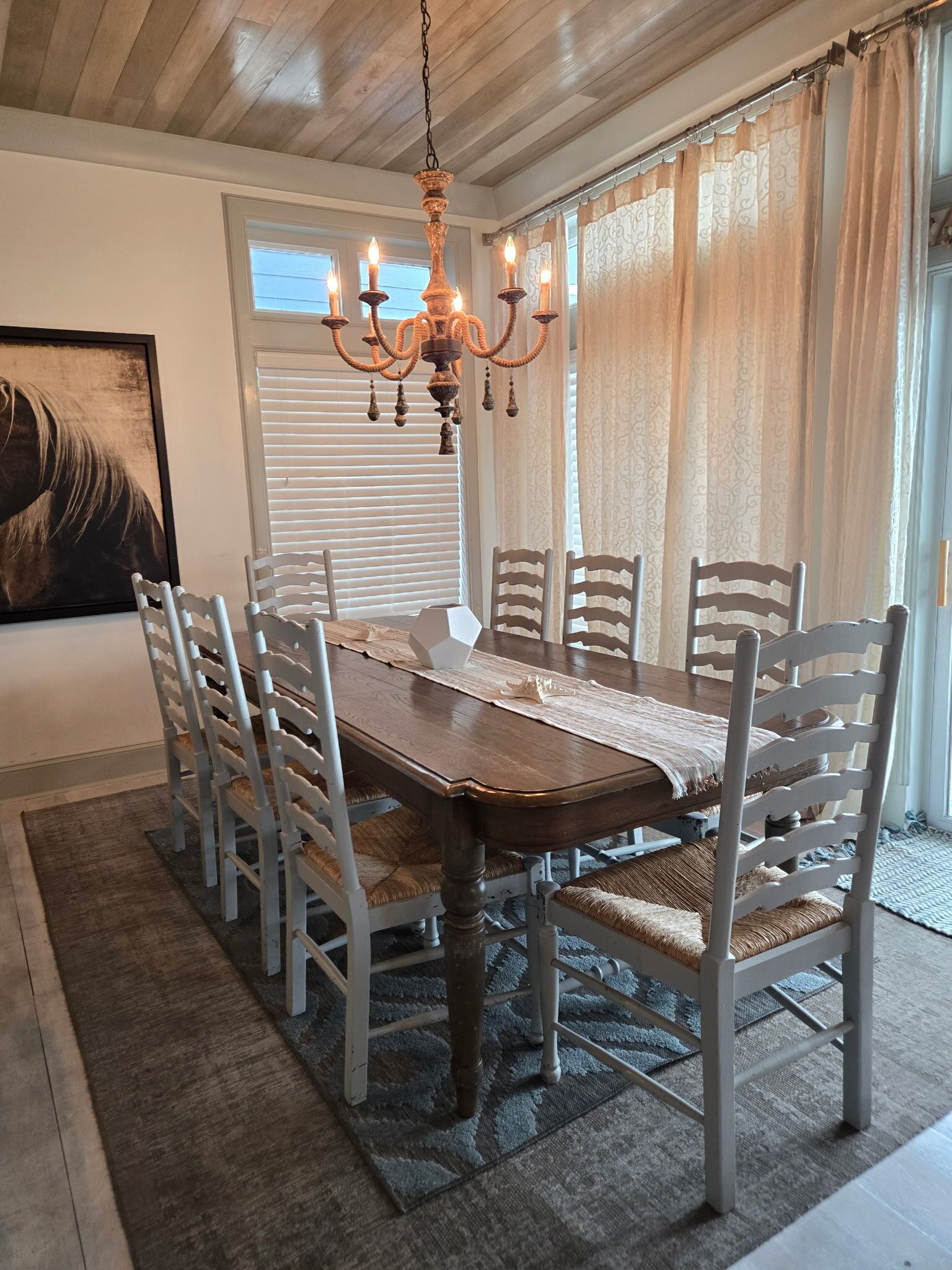 A dining room with a wooden table and white chairs, a chandelier, and a large window with curtains.