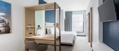 Una habitación de hotel con una cama, un escritorio con silla, un televisor y un ventanal grande.