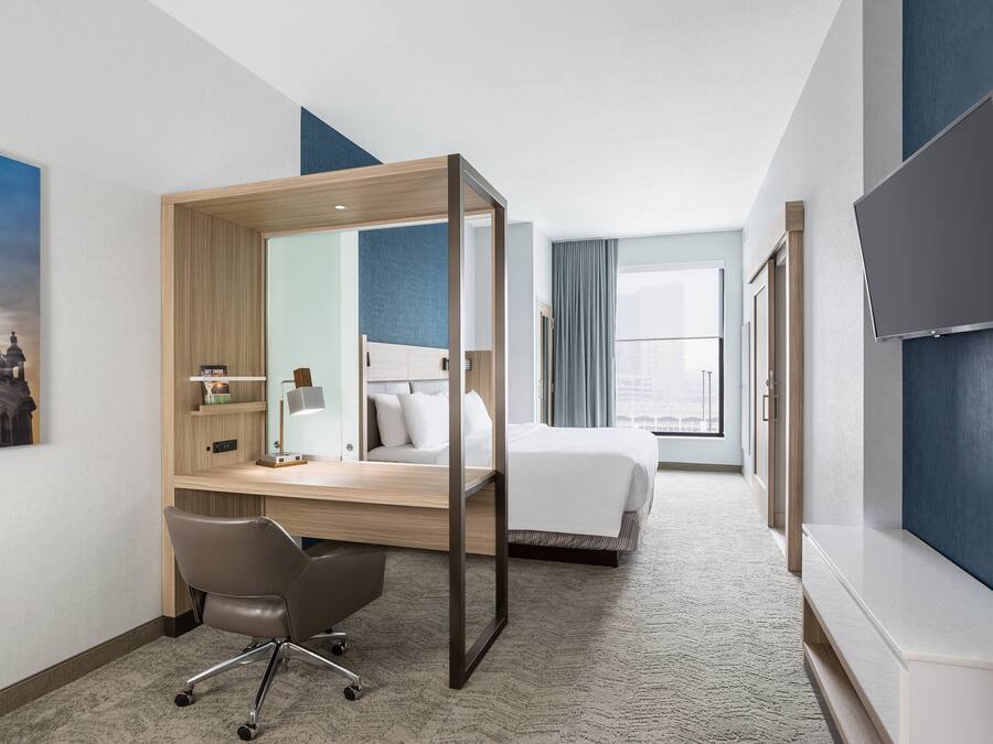 Una habitación de hotel con una cama, un escritorio con silla, un televisor y un ventanal grande.
