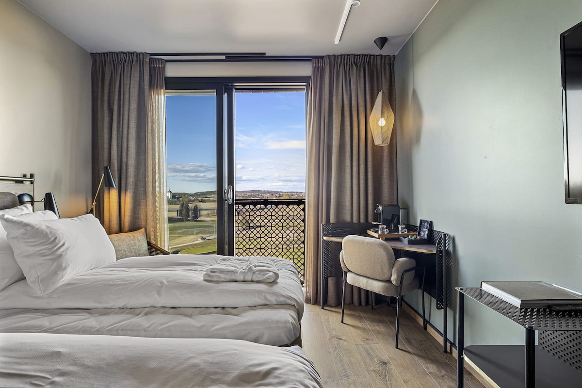 A hotel room with a bed, a desk, and a chair. There is a view of the outdoors through the window.