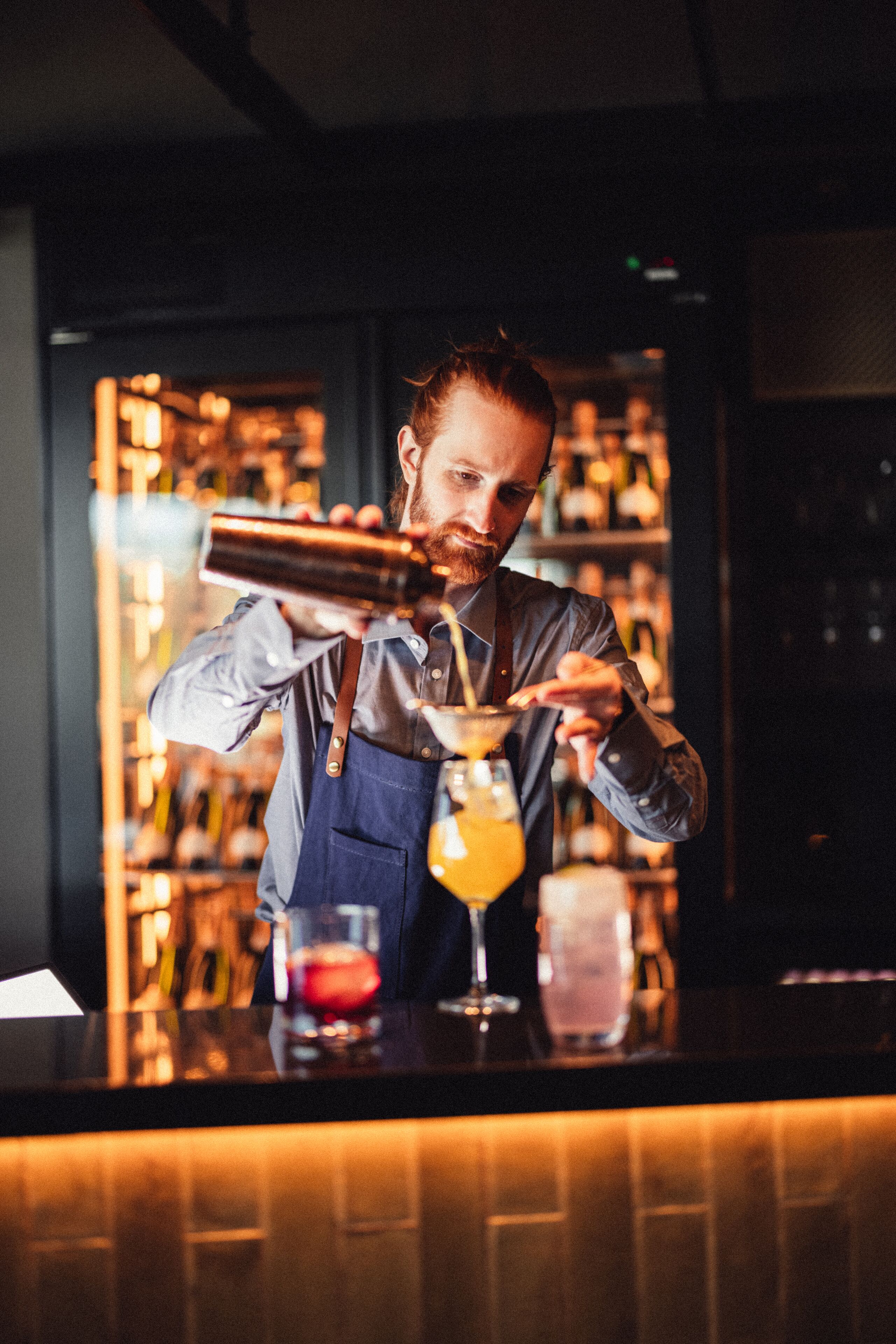 A bartender preparing a drink behind a bar with shelves of bottles and glasses.