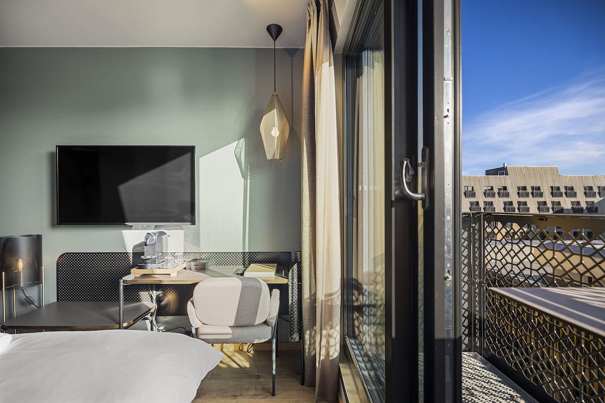 A hotel room with a bed, a desk, a chair, a TV, and a balcony with a view of buildings.