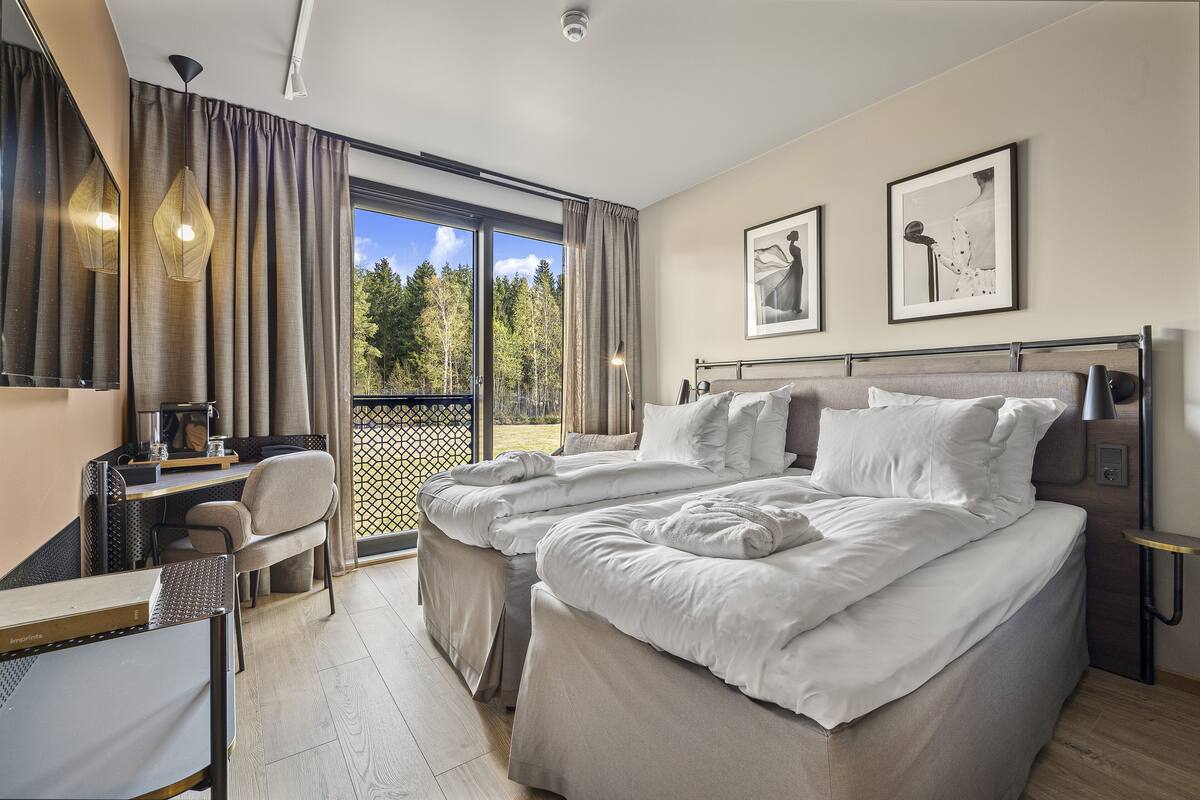 A bedroom with a large bed, a desk, a chair, and a balcony with a view of trees.