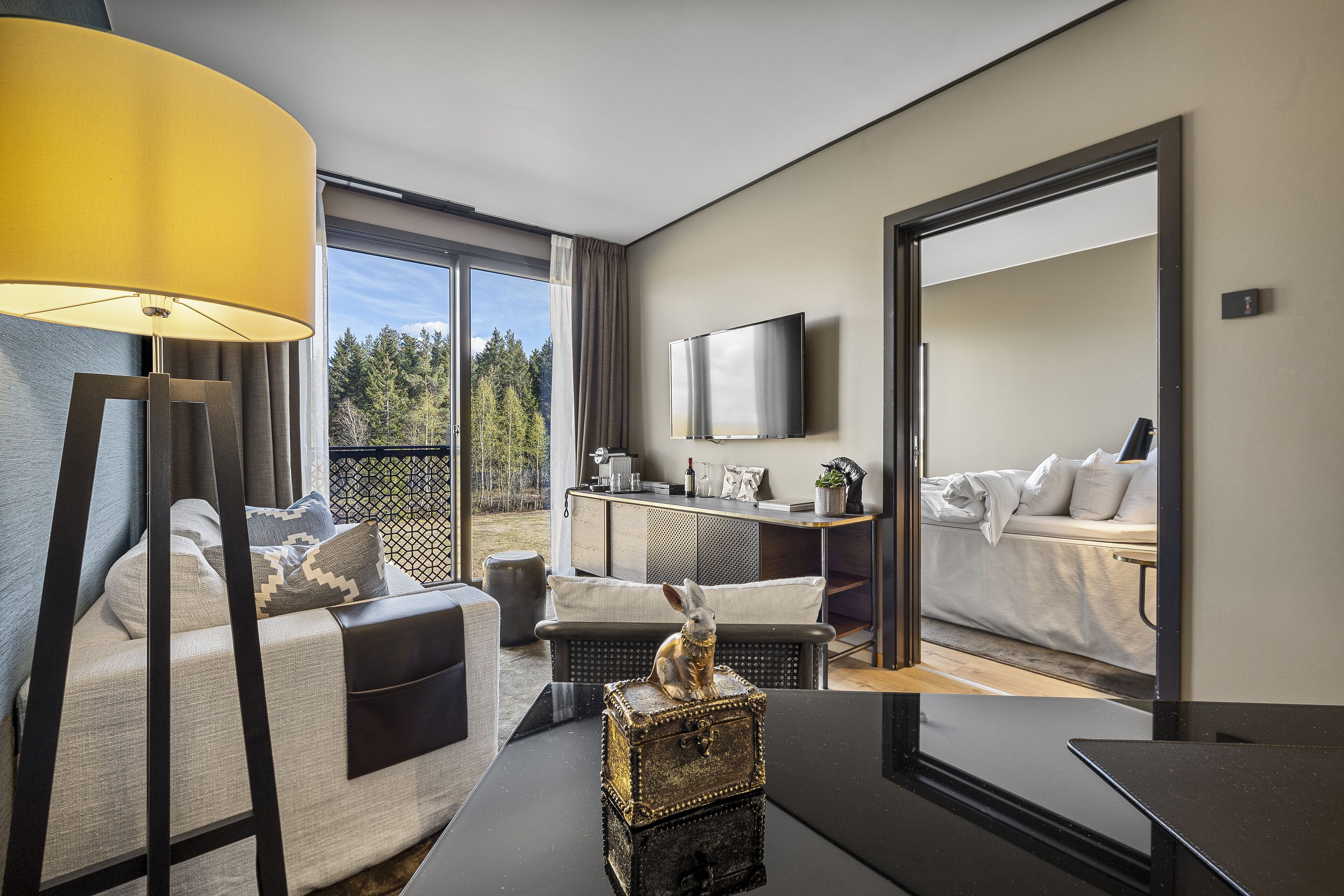 A modern hotel room with a sofa, a table with a lamp, a TV, and a view of the outdoors.