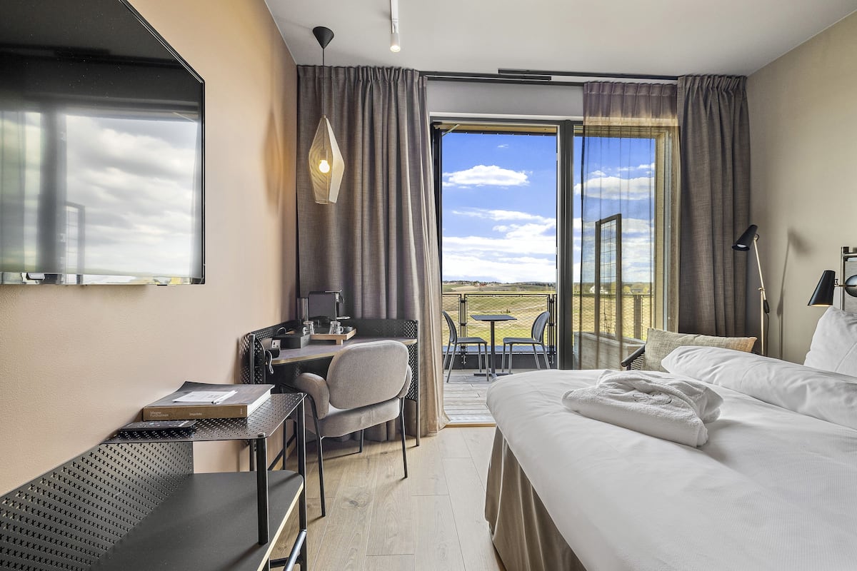 A hotel room with a bed, a desk, a chair, a small table, and a balcony with a view of the sky and clouds.
