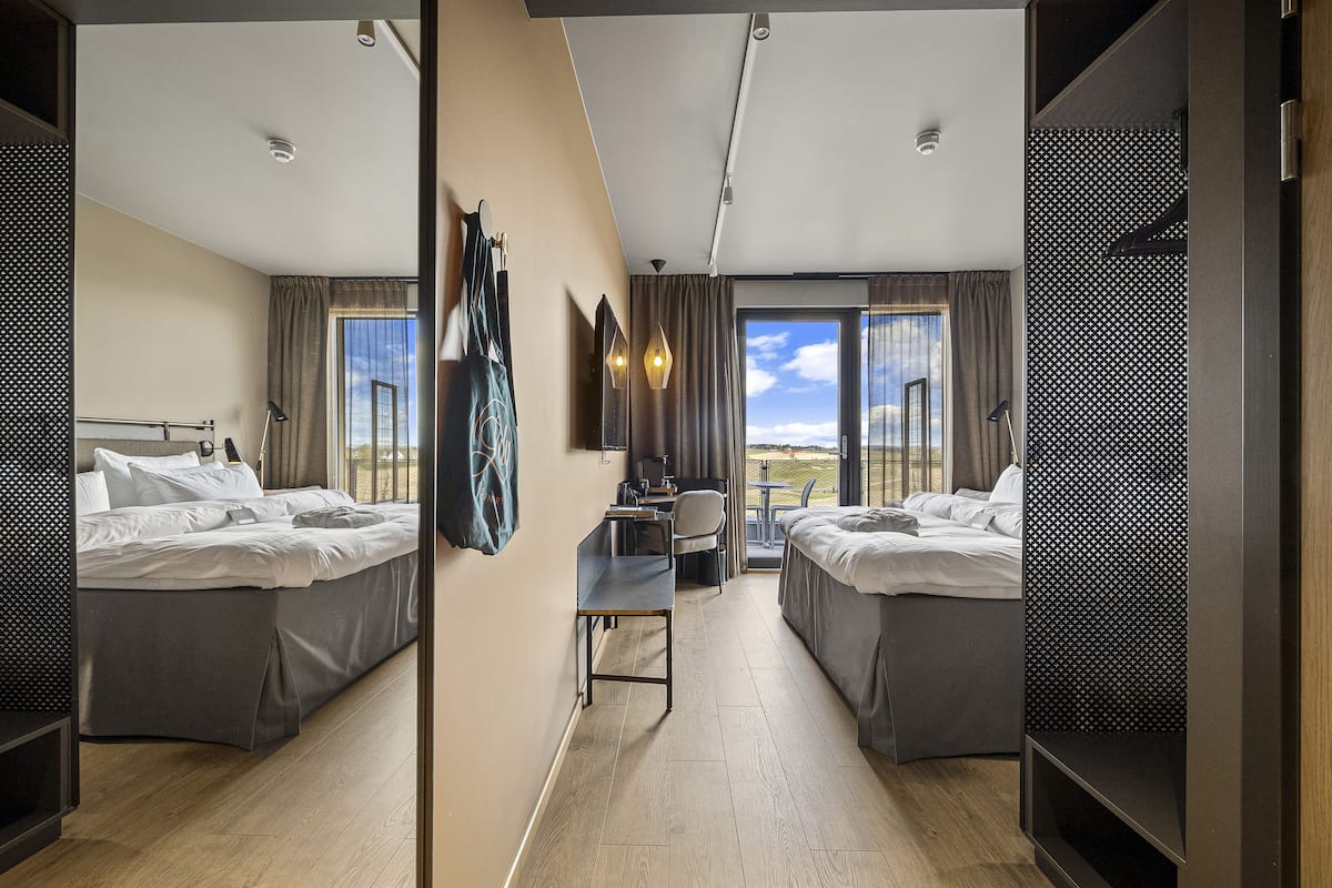 A modern hotel room with two beds, a desk, and a large window offering a view of the sky.