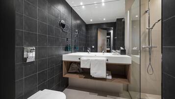 A modern bathroom with dark tiles, a large mirror, a sink with a towel rack, and a shower area.