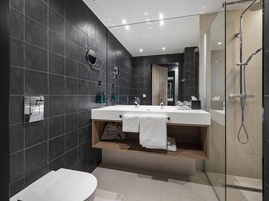 A modern bathroom with dark tiles, a large mirror, a sink with a towel rack, and a shower area.