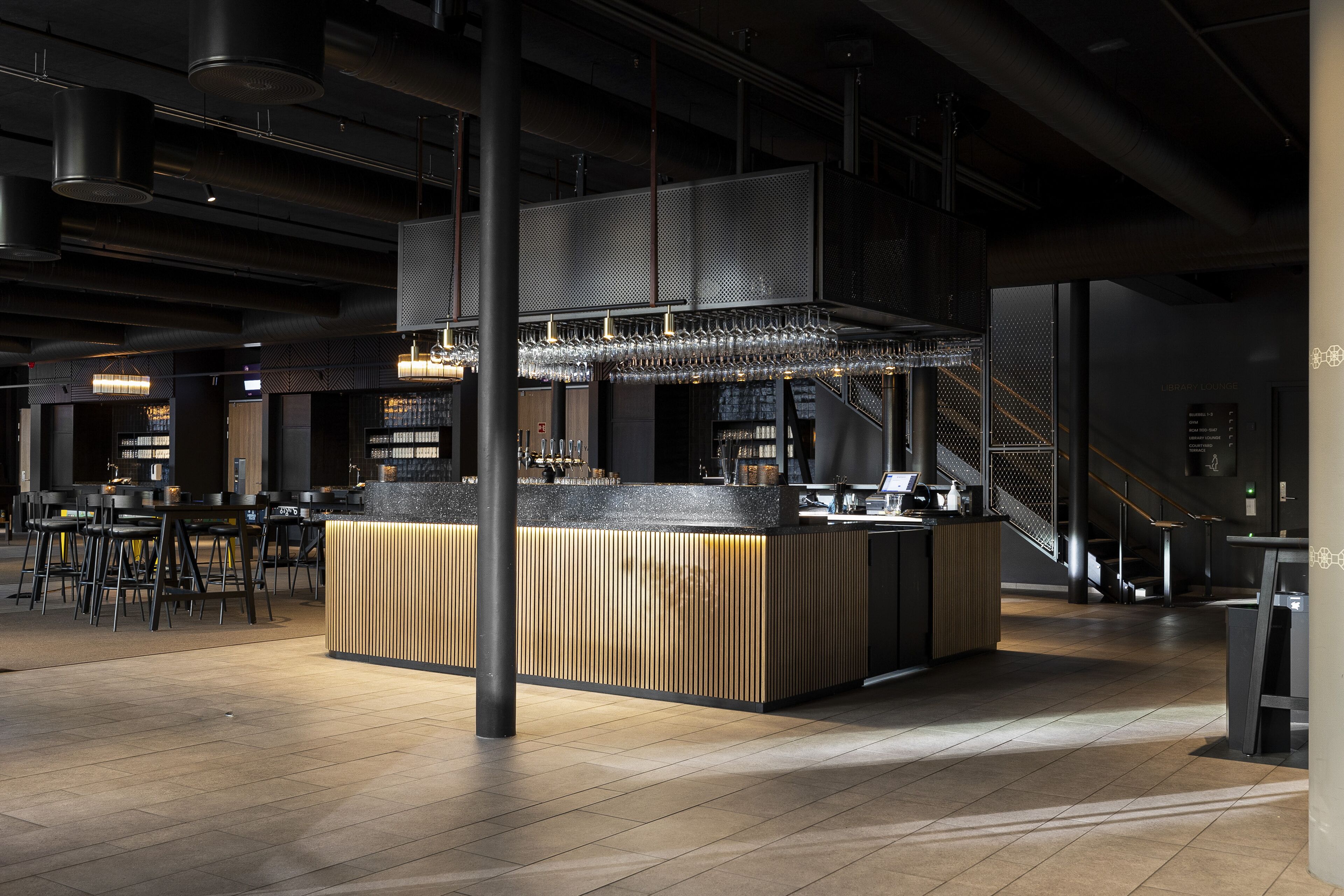 Un área de bar moderna con una barra central, diversas opciones de asientos y una escalera que sube a un nivel superior.