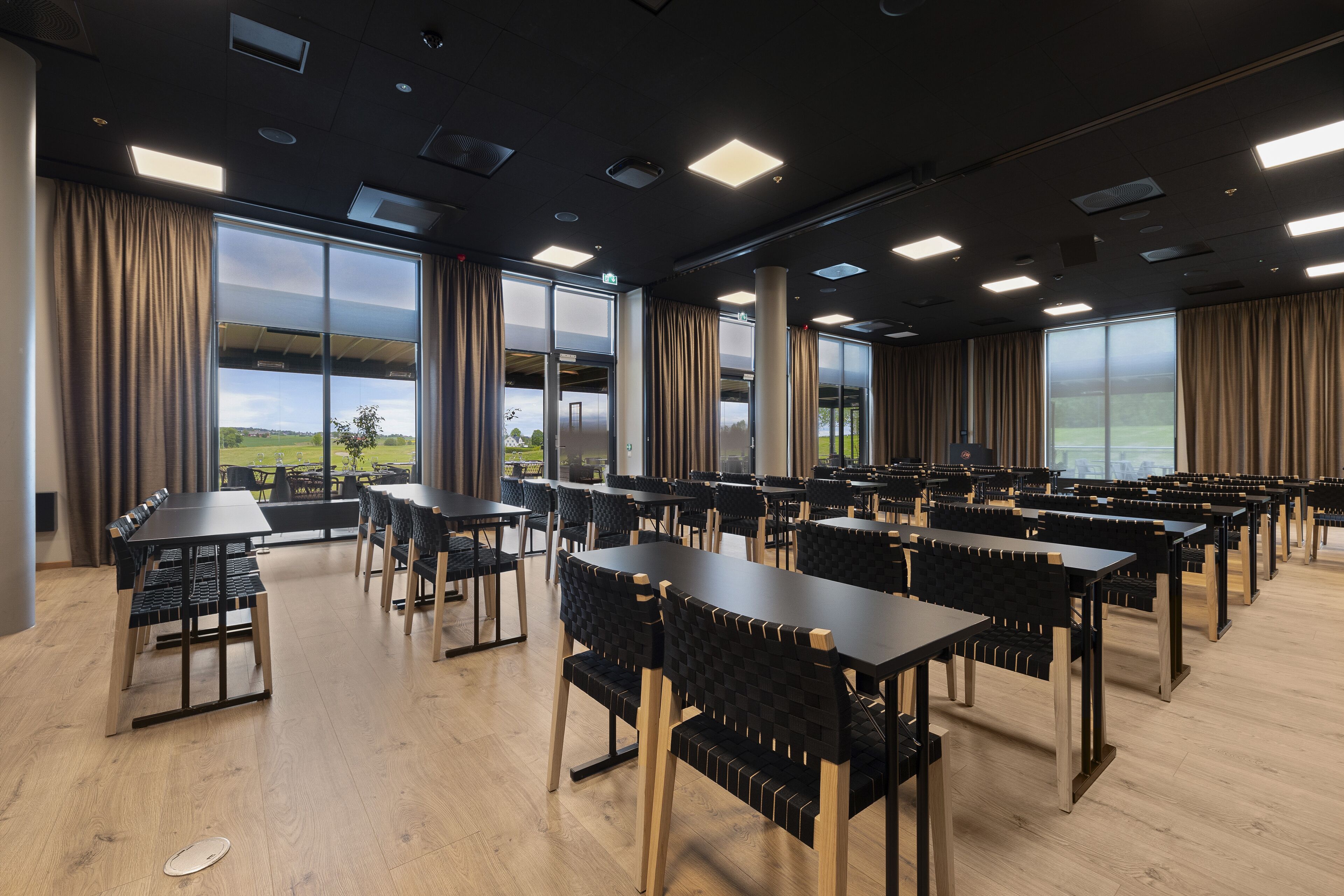Een ruime eetruimte met meerdere tafels en stoelen, grote ramen met gordijnen en uitzicht op de omgeving.