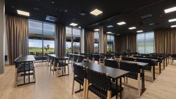Een ruime eetruimte met meerdere tafels en stoelen, grote ramen met gordijnen en uitzicht op de omgeving.