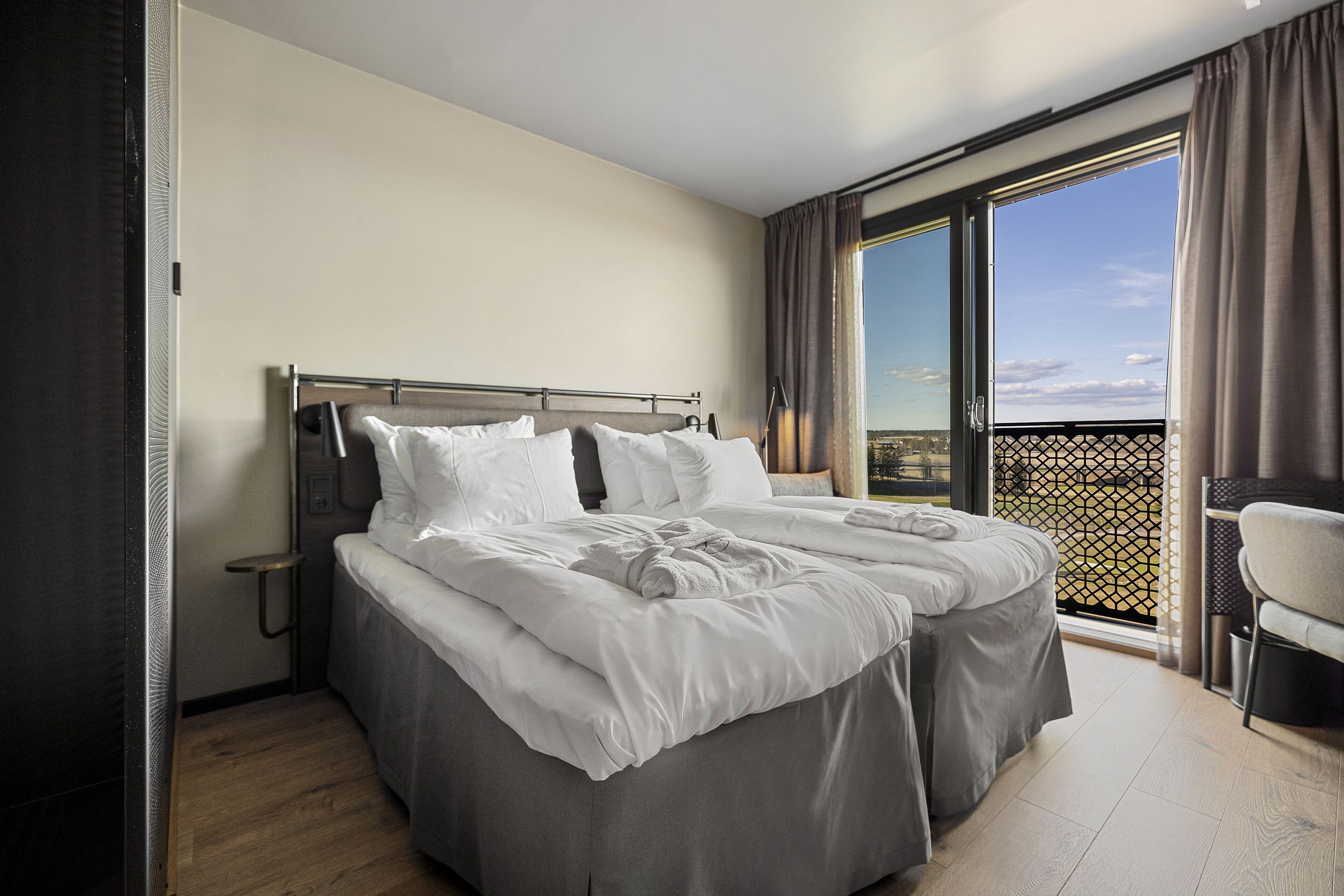 Ein Doppelbett mit weißen Bettwäsche, ein Nachttisch und Blick ins Freie durch ein großes Fenster.