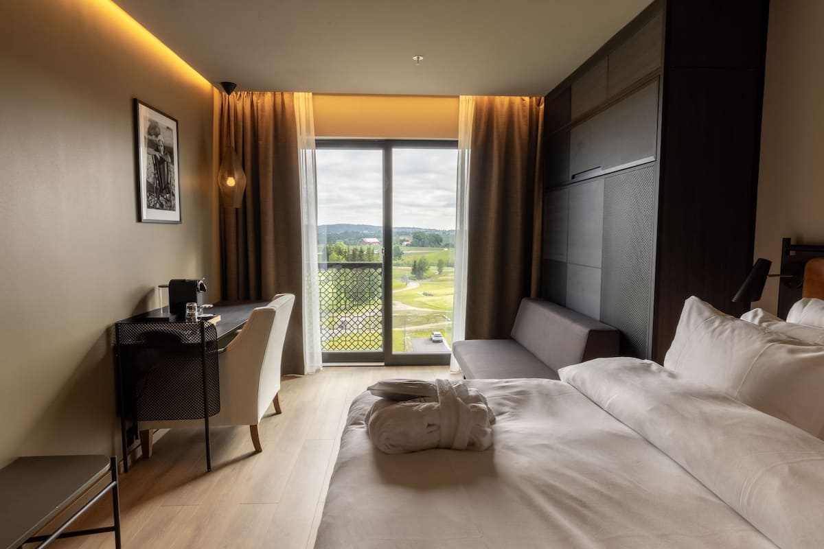A hotel room with a bed, a desk, a chair, a sofa, and a view of a golf course through a large window.