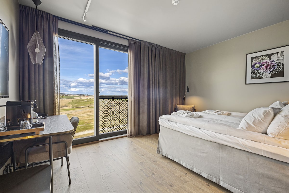 A bedroom with a bed, a desk with a computer, and a view of a golf course through a large window.