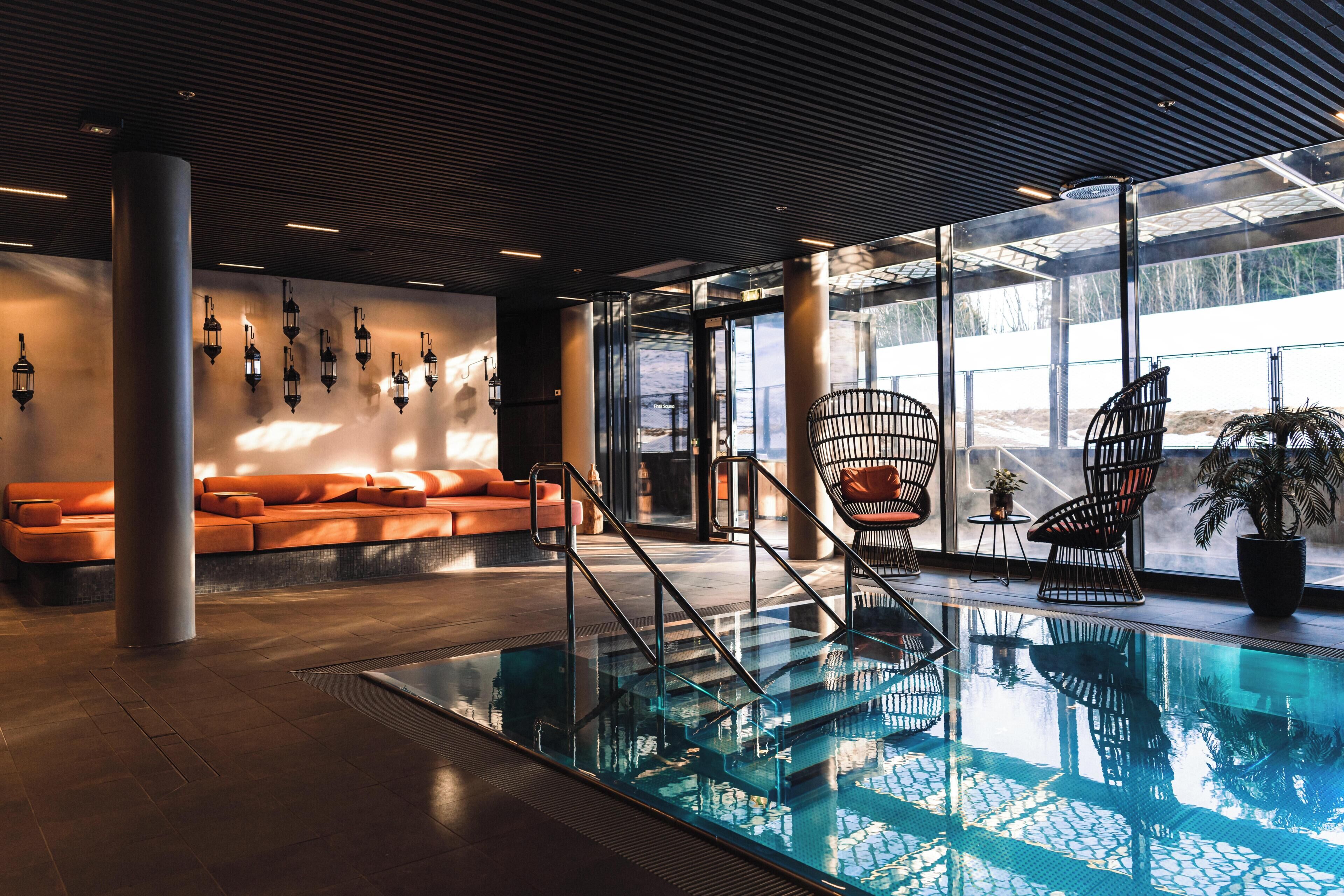Une piscine intérieure dotée de rampes en verre, un coin salon avec des canapés orange et une vue dégag ée grâce à de grandes fenêtres.