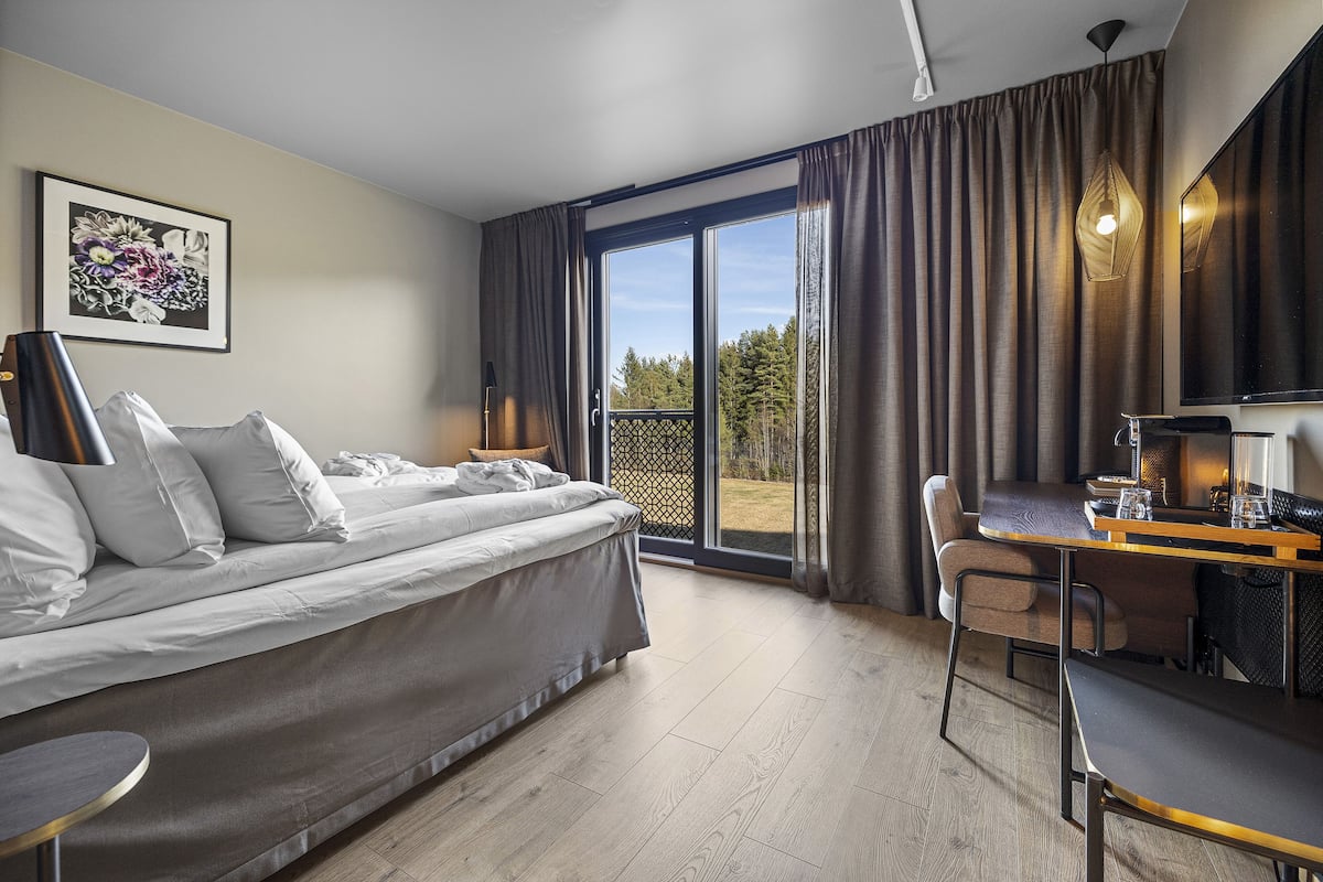 A modern hotel room with a bed, a desk, and a chair. There is a balcony with curtains and a view of trees.