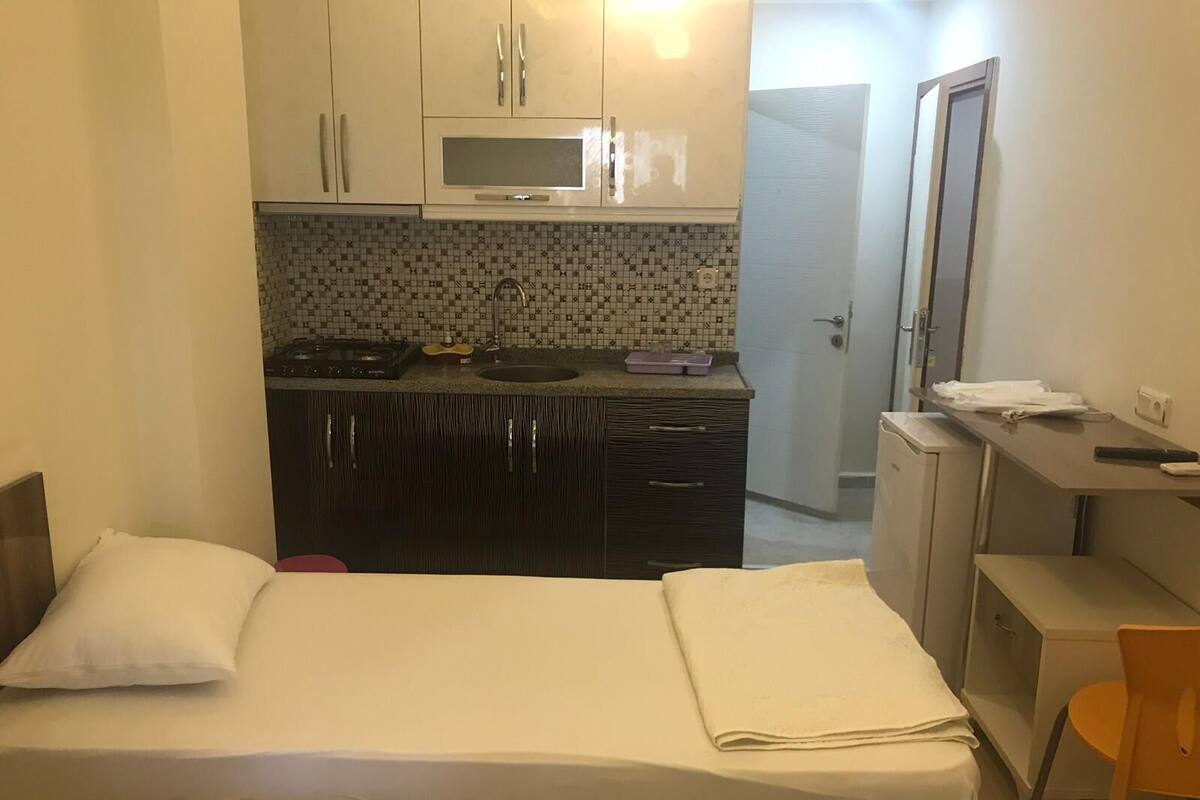 A compact hotel room with a bed, a small kitchenette, a desk, and a chair.