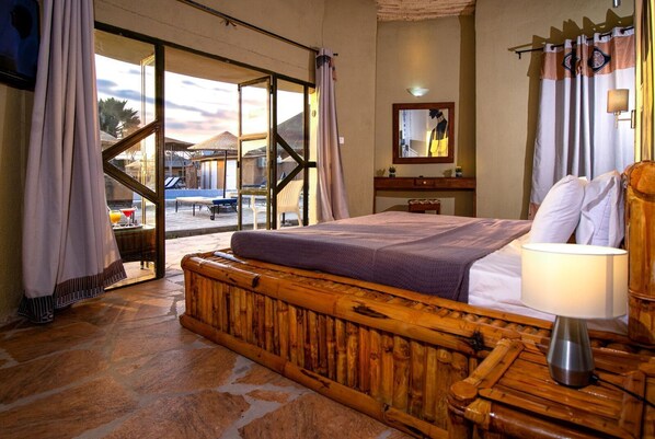A bedroom with a large bed, a wooden headboard, a nightstand, a lamp, and a view of the outside through a glass door.