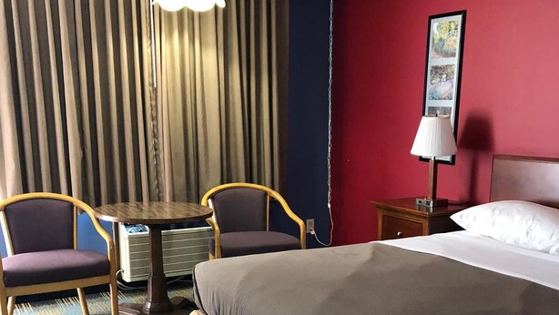 A hotel room with a bed, two chairs, a small table, a lamp, and a painting on the wall. - Rose Motel (David City)