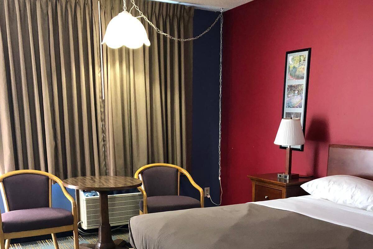 A hotel room with a bed, two chairs, a small table, a lamp, and a painting on the wall.