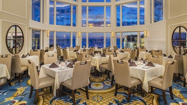 A large, well-lit dining area with multiple tables and chairs, large windows offering a view of the evening sky and buildings, and a patterned floor.