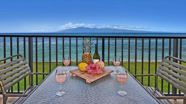 A table with a tropical fruit arrangement and wine glasses, overlooking a clear ocean and distant mountains.