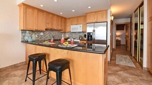A modern kitchen with wooden cabinets, a granite countertop, a stainless steel refrigerator, and two black bar stools.