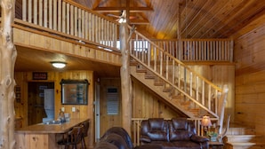 A wooden cabin interior with a dining area, a living room with a leather sofa, and a staircase. - Secluded Log Cabin, Mountain Top, Hot Tub, Pool Table, 5 Miles From Downtown (Hot Springs)