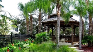 Ein Gazebo mit Holzdach und offenen Seiten, umgeben von üppigem Grün und Palmen.
