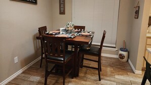 Un coin repas avec une table en bois dressée pour quatre personnes, entourée de chaises. On y trouve des objets décoratifs, un panneau mural sur lequel est écrit « Rassemblez-vous autour de la table », et un autre panneau sur lequel est écrit « À table ».