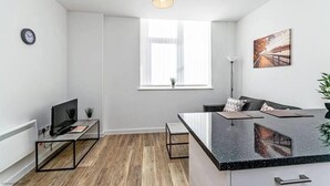 A modern living room with a wooden floor, a flat-screen TV, a coffee table, a sofa, a dining area, and a large window with blinds.