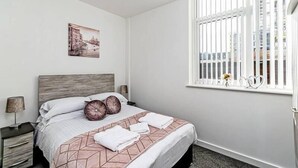 A bedroom with a bed, a nightstand, a lamp, a window with blinds, and a wall-mounted picture.